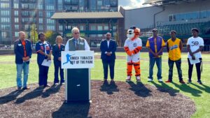Dean Aksu behind a podium at The Corner Ballpark speaking on oral health and the risks of tobacco use at a community awareness event.